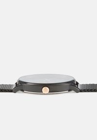 Black watch with a round face and mesh strap, featuring a clear glass cover and a gold-tone crown for adjustment. Minimalistic design.