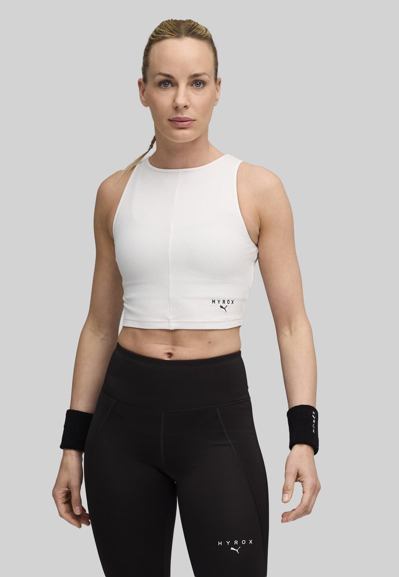 Hvid ribbet crop top med logo, parret med højtsiddende sorte leggings med logo. Sorte håndledsstøtter komplementerer outfittet.