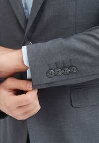 Manche de veste grise avec quatre boutons noirs. Poignet de chemise bleu clair visible en dessous. Texture de tissu lisse.