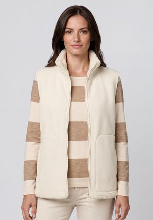 Woman wearing cream-colored fleece vest over beige and cream wide striped long sleeve shirt against plain light background.