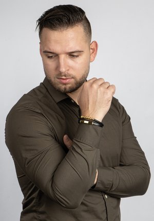 Braunes, tailliertes Langarmhemd aus strukturiertem Stoff mit Knöpfen. Trägt schwarze und goldene Armbänder am Handgelenk, die den Kontrast betonen.