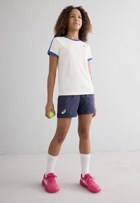 T-shirt atlético branco com detalhes azuis, combinado com calções azul-marinho. O modelo segura uma bola de ténis verde, vestindo meias brancas e sapatilhas de desporto cor-de-rosa.