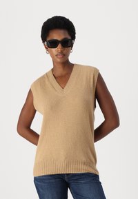 Beige ärmelloser Pullover mit V-Ausschnitt und geripptem Saum. Getragen über dunkelblauen Jeans; kombiniert mit schwarzen rechteckigen Sonnenbrillen.