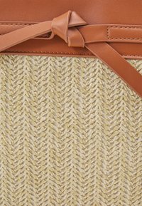 Wiklinowa torebka ze słomy z fakturowanym kremowym wzorem, z brązowym skórzanym paskiem górnym i detalem węzła. Kompaktowy, usztywniony kształt.