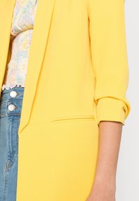 Žlutý blazer se strukturovaným designem, se zářezem na jedné kapse a vyhrnutými rukávy. Tkanina vypadá hladce a lehce.