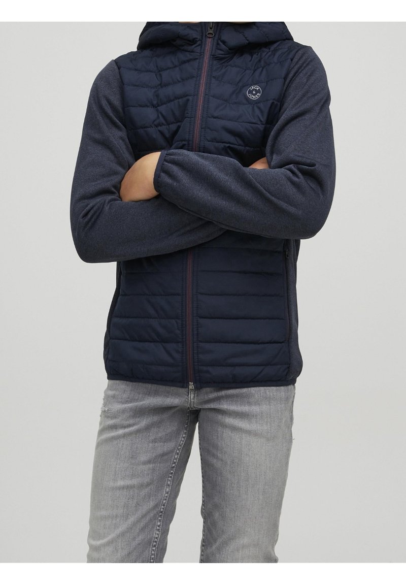 Veste bleu marine avec un devant matelassé, des manches en tissu gris, fermeture éclair et une capuche. Présente un patch logo sur la poitrine.