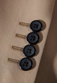 Beiger Stoff mit vier senkrecht angeordneten schwarzen Knöpfen, zeichnet sich durch eine glatte Textur und kontrastierende Nahtdetails aus.