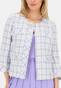 Biała i jasnoniebieska marynarka tweedowa z postrzępionymi brzegami, rękawami o długości trzy czwarte, dwiema przednimi kieszeniami oraz akcentem w postaci kwiatu blisko górnego zapięcia.