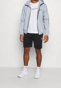 Mężczyzna w jasnoszarej kurtce z kapturem, białej koszulce z grafiką, czarnych szortach, białych skarpetkach Nike i białych sneakersach stoi na jednolitym tle.