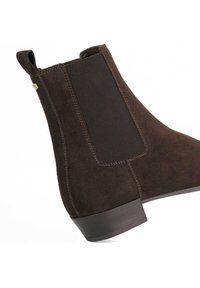 Bruine suede enkellaars met elastische zijkanten, een lage gestapelde hak en contrasterende stiksels langs de randen. Gladde textuur en gestroomlijnde vorm.