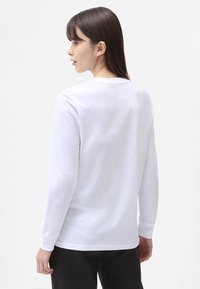 Hvid langærmet T-shirt, blødt bomuldsmateriale, rund halsudskæring, lige kant, synlige syninger, båret med sorte bukser.