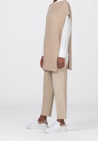 Beige Ripp-Pullover mit hohem Kragen und seitlichem Schlitz, getragen über einem weißen Hemd mit Knopfleiste, kombiniert mit beigen Hosen und weißen Turnschuhen.