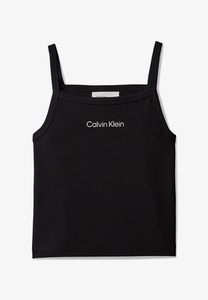 Dziewczyna z długimi brązowymi włosami nosi czarny top Calvin Klein, niebieskie dżinsy i czarne sandały, stoi z jedną ręką w kieszeni.