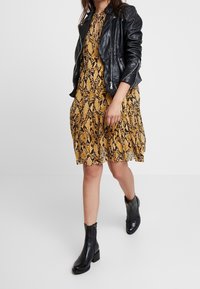 Černá kožená bikerská bunda přes žluté šaty s hadím vzorem a rozšířenou sukní, doplněná černými kotníkovými botami.