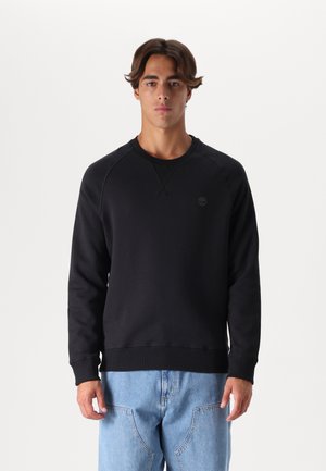 Zwarte crewneck sweatshirt met ribgebreide mouwen en zoom, voorzien van een reliëflogo op de borst. Gecombineerd met lichtblauwe spijkerbroek.