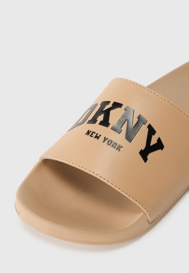 Sandalia deslizante marrón con una tira ancha que presenta el logo en negro "DKNY NEW YORK", material sintético suave, punta redondeada y un brillo sutil.