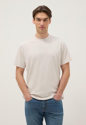 Joven de pie con las manos en los bolsillos, vistiendo una camiseta blanca lisa y jeans azules, sobre un fondo neutro.