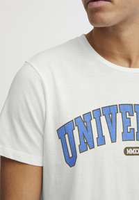 Balts kokvilnas T-krekls ar apaļu kaklu, uz krūtīm ar zilu un brūnu "UNIVERSITY" teksta grafiku, īsām piedurknēm un mīkstu tekstūru.