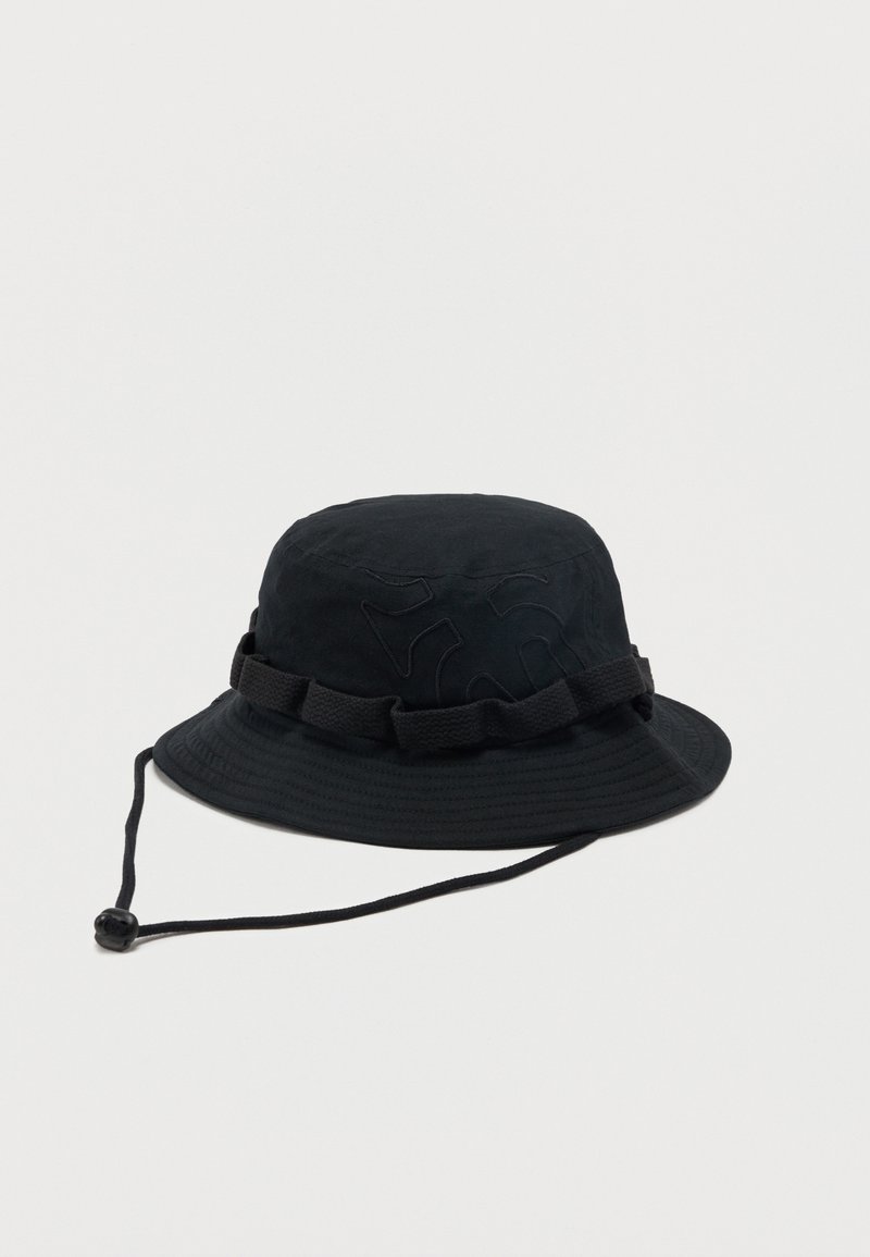 Sombrero tipo bucket negro con ala ancha, correa ajustable para la barbilla y detalles de lazo de tela alrededor de la corona, sobre un fondo liso.