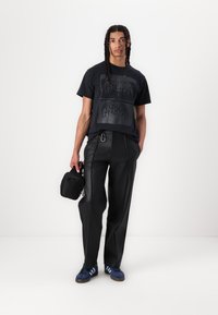 Black graphic t-shirt with a textured design, black faux leather pants, and blue sneakers. Small black bag held in left hand.