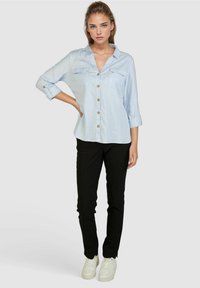 Helleblaue Button-down-Hemd mit hochgekrempelten Ärmeln, zwei Brusttaschen, getragen mit schwarzen Hosen und weißen Sneakern. Glatter Stoff und taillierte Passform.