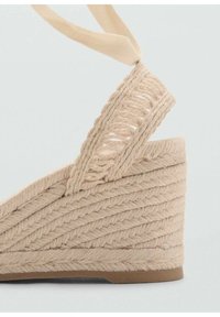 Beige wedge sandal with woven jute texture, featuring a thick, braided strap and a flat rubber sole. Accent of soft fabric ties.