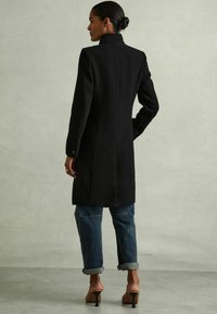 Abrigo negro, entallado, con un alto cuello y mangas largas. Presenta un corte recto y una textura sólida, combinado con vaqueros azules con puños y zapatos de tacón color nude.