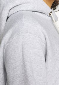 Gros plan d'une personne portant un sweat à capuche gris clair avec un cordon blanc et des coutures visibles aux épaules.