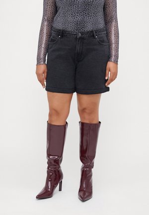 Mujer lleva shorts vaqueros negros con bajo vuelto, top de manga larga transparente con estampado de leopardo y botas de tacón brillantes hasta la rodilla en color burdeos oscuro.