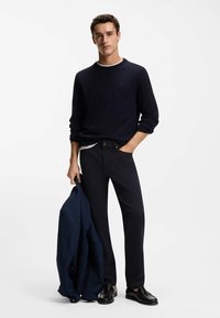 Granatowy sweter z dzianiny, biały kołnierzyk, ciemne spodnie, trzyma ciemną kurtkę i nosi czarne buty wsuwane. Proste, czyste linie i klasyczne kolory.