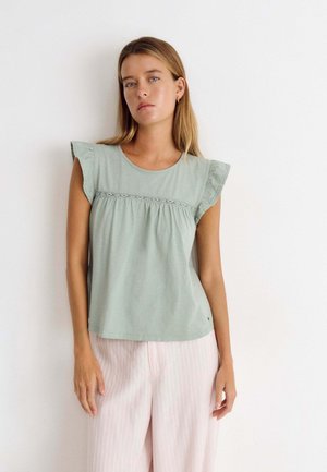 Mujer con cabello rubio liso que lleva una camiseta sin mangas verde claro con volantes en los hombros y pantalones anchos a rayas en rosa claro.