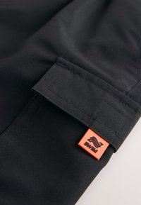 Sort stof med en glat tekstur har en foldet manchet og en orange mærkeetiket med en logo.