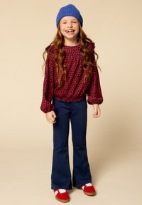 Rode blouse met bloemenpatroon en pofmouwen, gecombineerd met marineblauwe flared broeken. De outfit wordt aangevuld met rode schoenen en een blauwe gebreide muts.