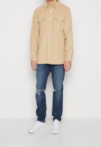 Beige corduroy overhemd met lange mouwen en twee borstzakken, gecombineerd met blauwe distressed jeans en witte sneakers. Klassiek knoopsluitingsontwerp.