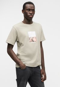 Ανοιχτό μπεζ βαμβακερό μπλουζάκι με ορθογώνιο γραφικό από βράχια και ουρανό, με λευκή λεπτομέρεια κειμένου. Το μοντέλο φορά σκούρο γκρι τζιν.