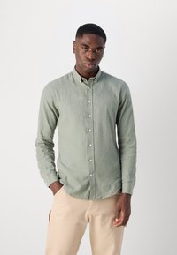 Cămașă lungă cu mâneci din liniș verde deschis, cu guler cu nasturi, fabricată din in, având nasturi pe față și o croială adaptată, asortată cu pantaloni bej.