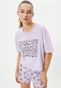 T-shirt di cotone viola chiaro con la scritta "DONUT WAKE ME UP"; abbinata a pantaloni corti coordinati con motivi di ciambelle e tazzine di caffè.