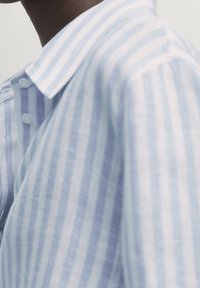 Camicia a righe azzurro chiaro e bianco con colletto abbottonato, caratterizzata da un tessuto texturizzato e bottoni visibili lungo la patta.