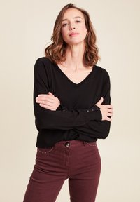 Schwarzes Langarmoberteil mit V-Ausschnitt und geknöpften Manschetten, kombiniert mit bordeauxroten Jeans mit dekorativer Naht und tailliertem Schnitt.