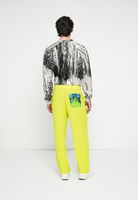 Langærmet skjorte med et sort og hvidt abstrakt mønster, kombineret med lyse neon gule bukser med en blå og grøn lomme detalje.