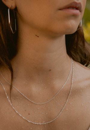 Three delicate silver chains layered around the neck, with water droplets on skin, and a large silver hoop earring in the ear.