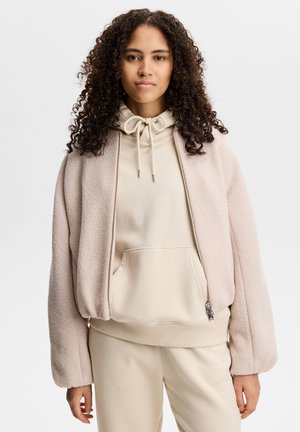 Jonge vrouw met krullend haar draagt een lichtbeige hoodie onder een zachte roze jas met rits en bijpassende lichtbeige broek.