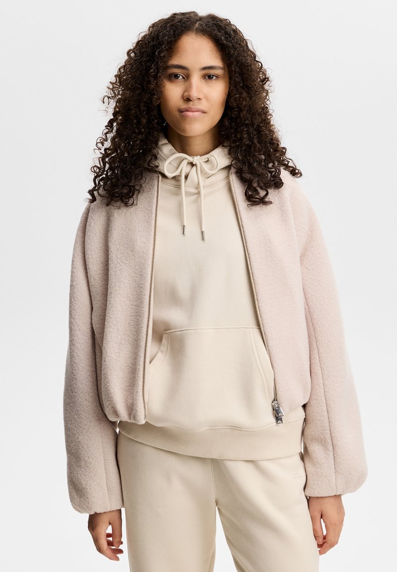Young woman with curly hair wearing a light beige hoodie under a soft pink zip-up jacket and matching light beige pants.