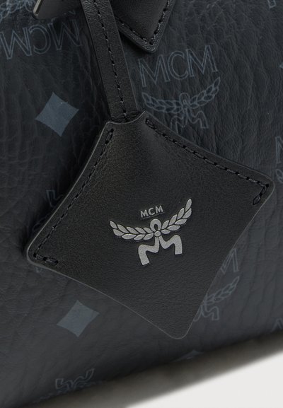 Melna ādas birka ar MCM sudraba logo, piestiprināta pie teksturētas melnas ādas somas ar atkārtotiem MCM logotipiem un dimanta formām.
