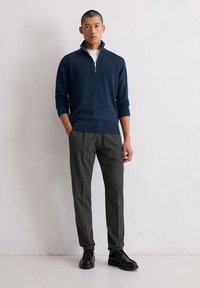 Marineblauer Zip-Kragen-Pullover aus strukturiertem Strick, kombiniert mit maßgeschneiderten grauen Hosen und schwarzen Schnürschuhen, stehend vor einem neutralen Hintergrund.