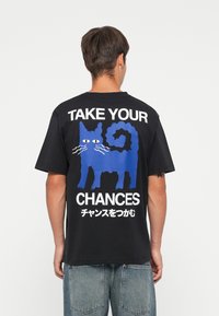 Zwarte katoenen t-shirt met een blauwe kattenafbeelding en witte tekst "NEEM JE KANSEN" met Japanse karakters op de rug. Korte mouwen, relaxte pasvorm.