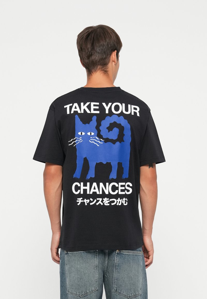Zwarte katoenen t-shirt met een blauwe kattenafbeelding en witte tekst "NEEM JE KANSEN" met Japanse karakters op de rug. Korte mouwen, relaxte pasvorm.