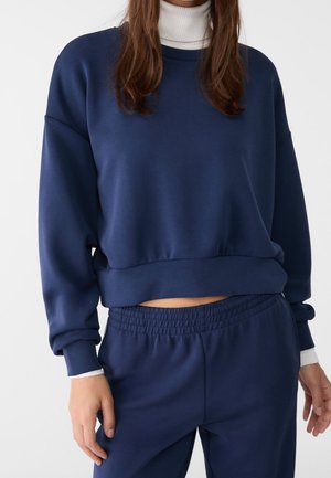 Kvinde iført en mørkeblå cropped sweatshirt og matchende bukser med elastisk talje over en hvid rullekrave, stående foran en ensfarvet baggrund.