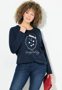 T-shirt a maniche lunghe color navy con una grafica di un leone bianco con un cappello da Babbo Natale e la scritta "Buone Feste". Il modello tiene in mano una borsa trapuntata rossa.