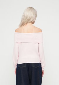 Gaiši rozā džemperis ar nokratītu plecu dizainu, ar ribota auduma tekstūru apkārt kakla un piedurknēm, apvienots ar tumši ziliem platām kājām džinsiem.